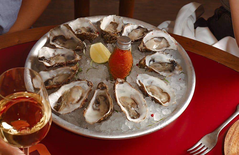 A circular tray of oysters on a bed of ice, garnished with a lemon and a bottle of sriracha.